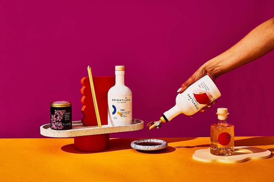 Still life shot of Brightland olive oils and vinegars on a platform in front of a fuchsia and orange background with an arm pouring oil.