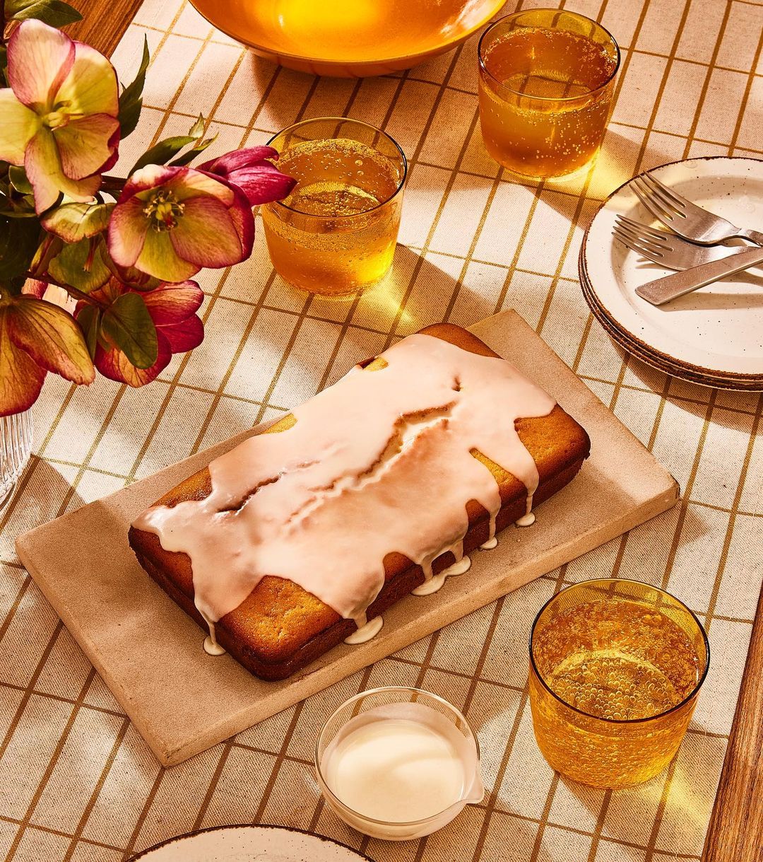 Overhead shot of a loaf cake with white glaze on a wooden board, glasses of orange liquid, flowers, and plates on a plaid tablecloth.