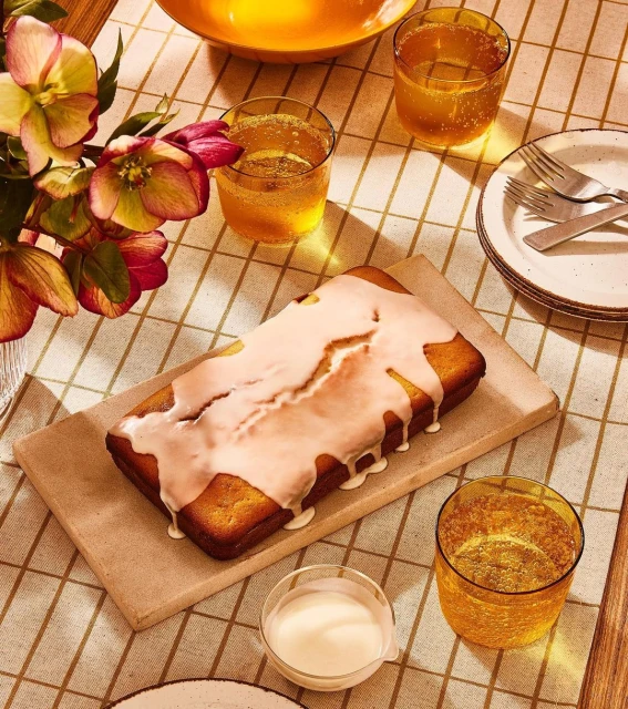 Overhead shot of a loaf cake with white glaze on a wooden board, glasses of orange liquid, flowers, and plates on a plaid tablecloth.