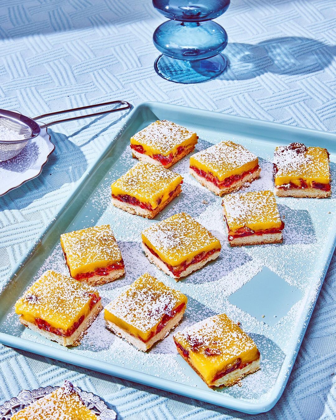 Overhead shot of a tray of square dessert bars topped with powdered sugar, featuring layers of yellow curd and red fruit filling.