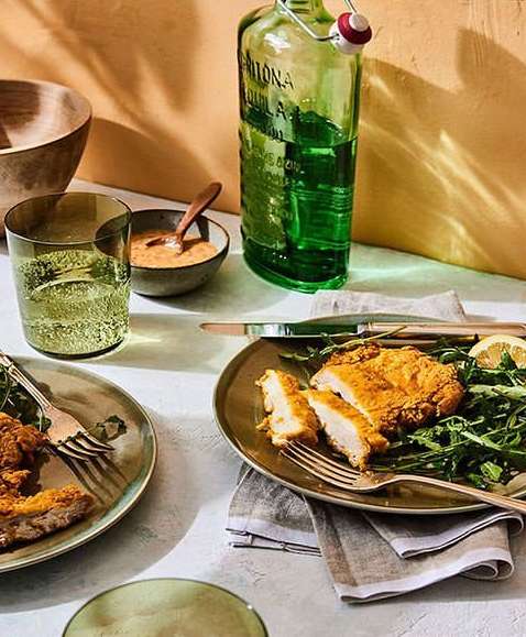 Overhead shot of two plates with golden fried cutlets, greens, lemon, drinks, and dipping sauce arranged on a white-grey table.