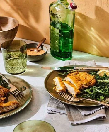 Overhead shot of two plates with golden fried cutlets, greens, lemon, drinks, and dipping sauce arranged on a white-grey table.