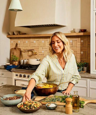 Woman prepares meal with vegetables and herbs on marble countertop in neutral kitchen, lit by natural light and modern pendant.