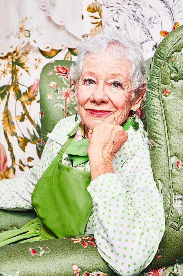 Elderly woman with gray hair sits in a green floral armchair, wearing a polka dot blouse and apron, hand to chin, smiling indoors.