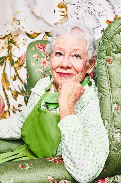 Elderly woman with gray hair sits in a green floral armchair, wearing a polka dot blouse and apron, hand to chin, smiling indoors.