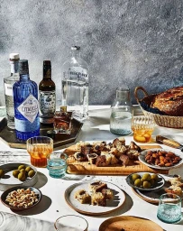 Cocktail bottles, glassware, olives, nuts, and sliced bread arranged on a table against a textured gray wall.