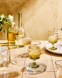 Green glassware filled with ice and amber liquid on a tiled tabletop with wine bottles and white flowers.