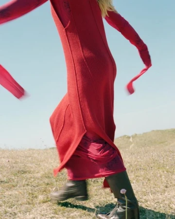 Model in a long red dress and dark boots walking across a grassy outdoor landscape under a clear sky.