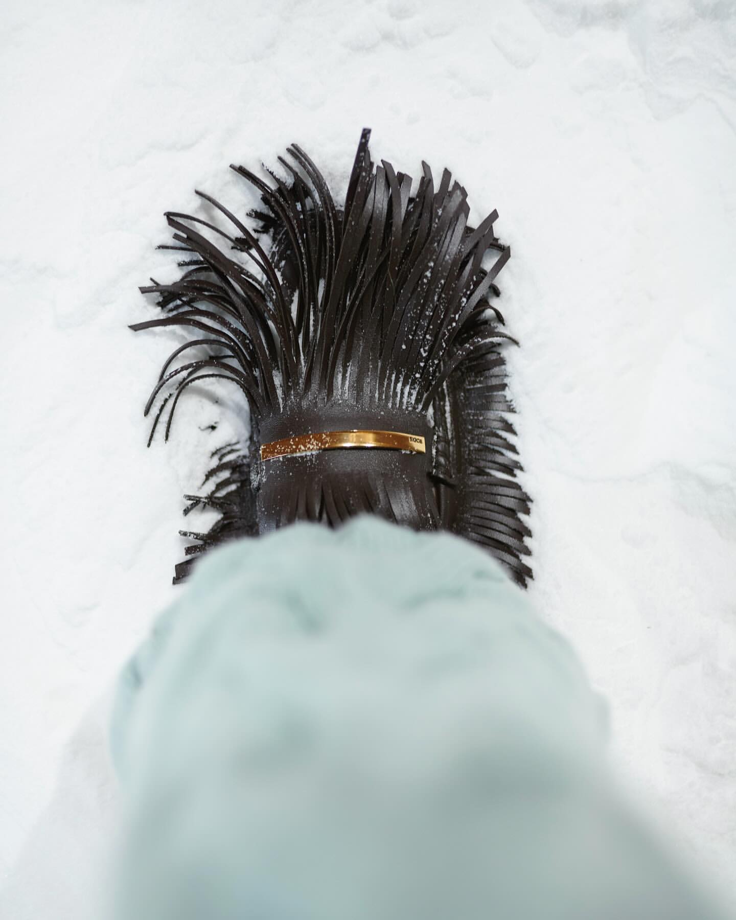 Overhead shot of a black leather fringe handbag with a gold bar detail placed on snow. The lower portion of a coat sleeve is visible.