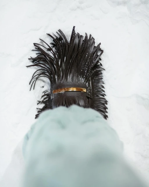 Overhead shot of a black leather fringe handbag with a gold bar detail placed on snow. The lower portion of a coat sleeve is visible.