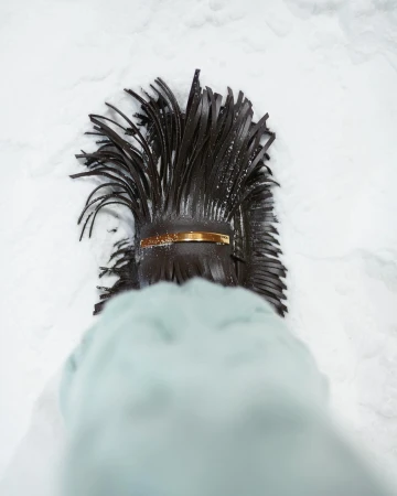 Overhead shot of a black leather fringe handbag with a gold bar detail placed on snow. The lower portion of a coat sleeve is visible.