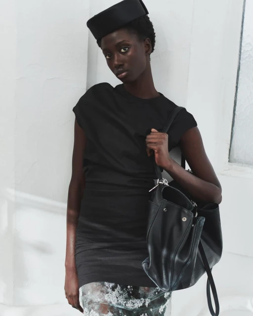 Elegant studio shot of a Black woman wearing a black dress and hat, carrying a leather handbag, against a white wall, fashion.