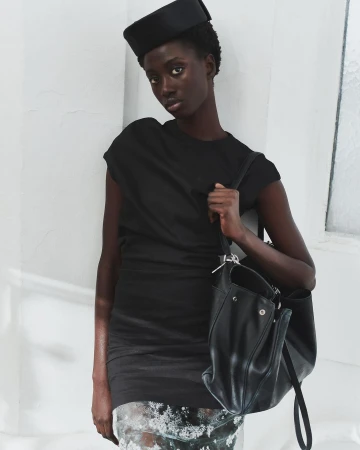 Elegant studio shot of a Black woman wearing a black dress and hat, carrying a leather handbag, against a white wall, fashion.