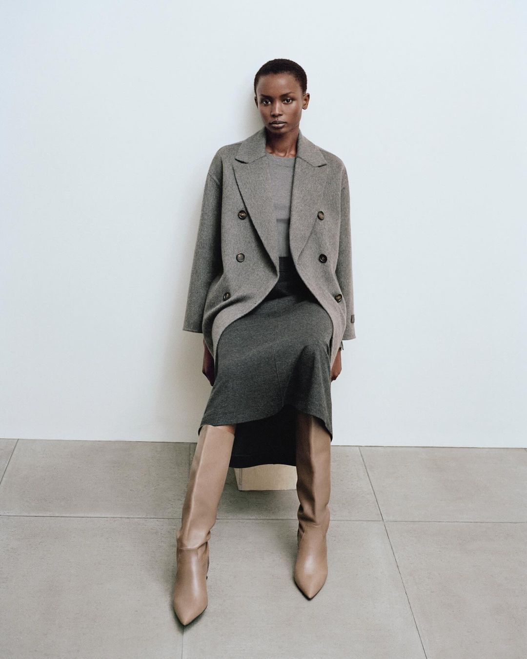 Woman in a grey double-breasted coat and dress sits indoors with beige boots in a fashion editorial, showcasing neutral tones and classic style.