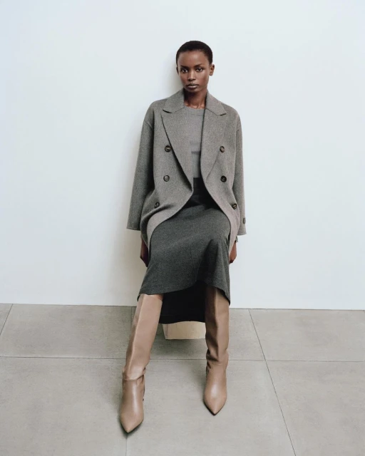 Woman in a grey double-breasted coat and dress sits indoors with beige boots in a fashion editorial, showcasing neutral tones and classic style.