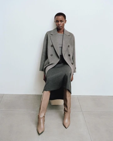 Woman in a grey double-breasted coat and dress sits indoors with beige boots in a fashion editorial, showcasing neutral tones and classic style.