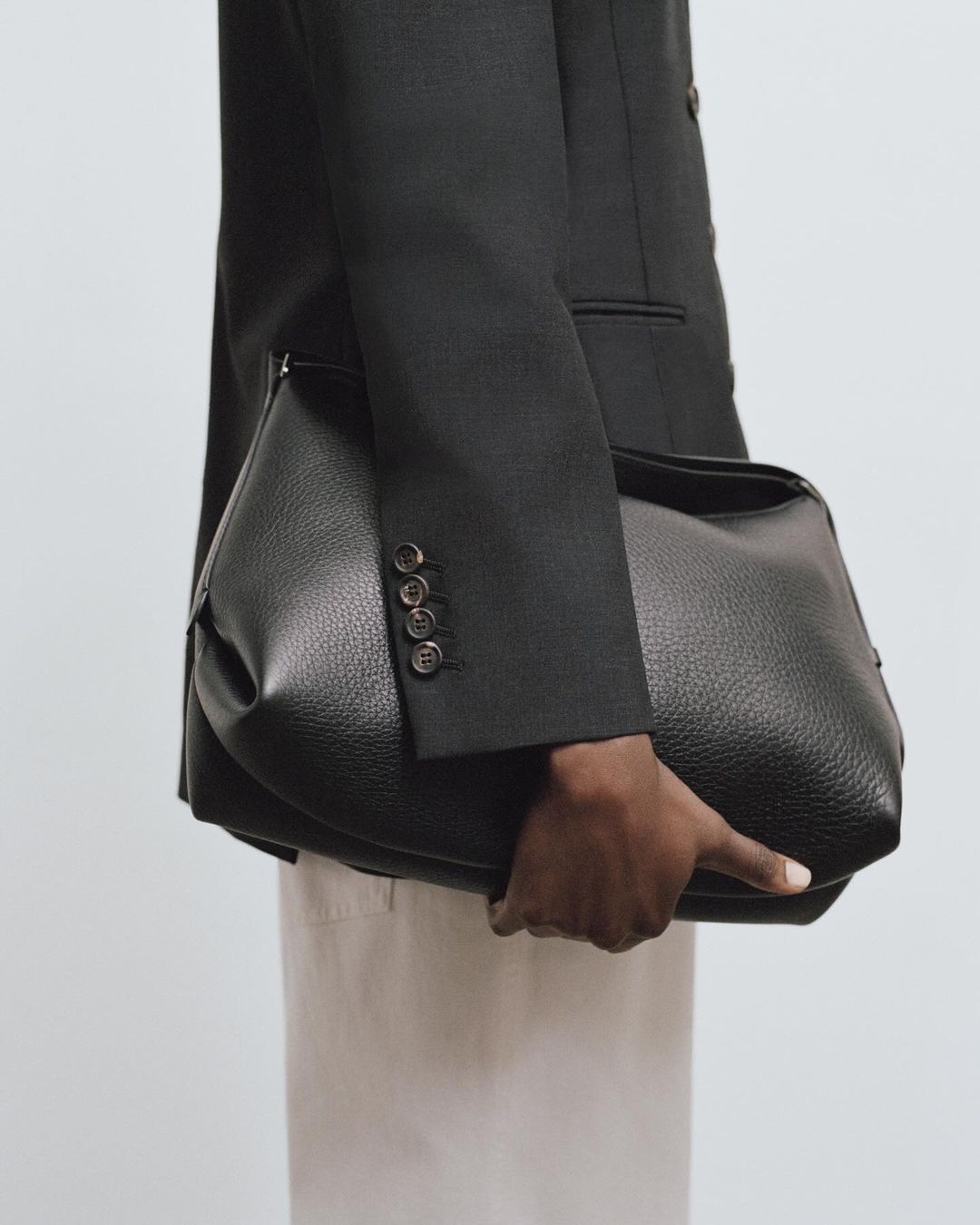 Close-up of a person holding a large black leather handbag wearing a dark blazer and light trousers against a neutral background.