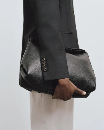 Close-up of a person holding a large black leather handbag wearing a dark blazer and light trousers against a neutral background.