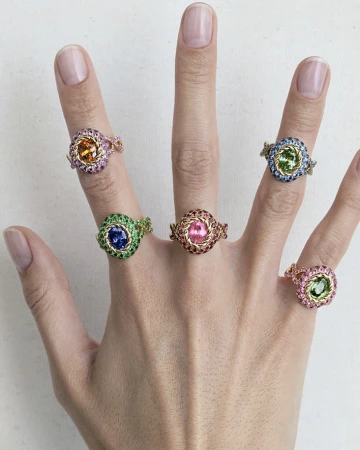 Close-up of a hand wearing five intricate jeweled rings with colorful gemstones on every finger by Aude Le Barbey