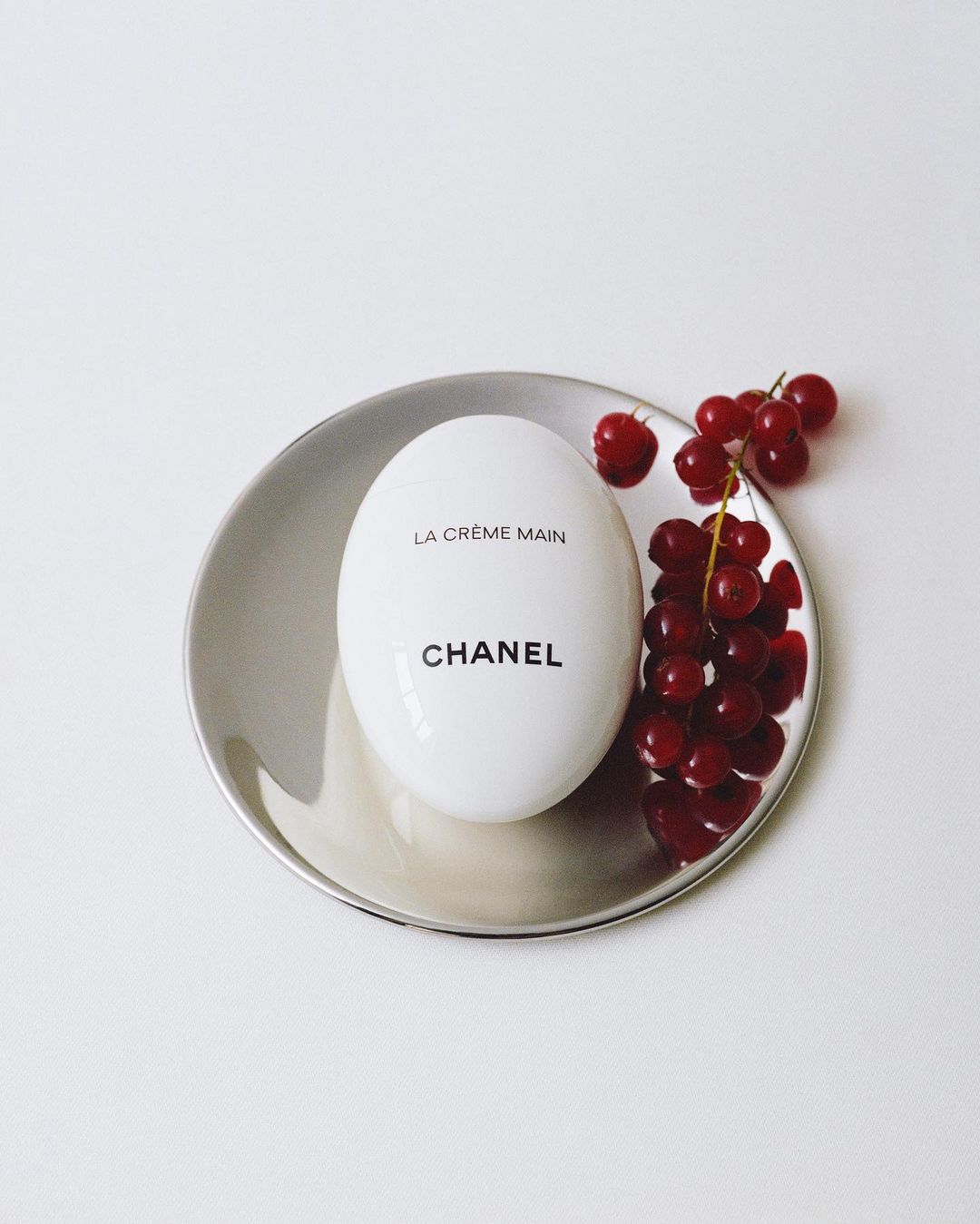 Luxury beauty product still life: Chanel La Crème Main hand cream on a silver dish with fresh red currants, minimalist aesthetic.