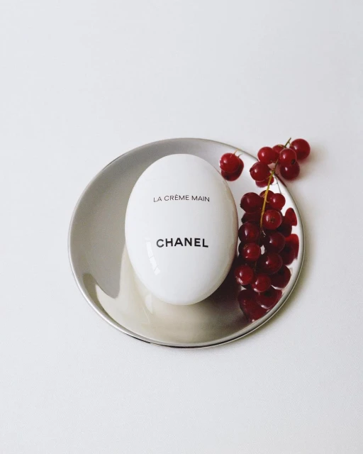 Luxury beauty product still life: Chanel La Crème Main hand cream on a silver dish with fresh red currants, minimalist aesthetic.