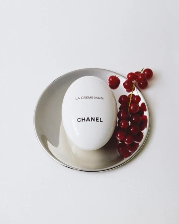 Luxury beauty product still life: Chanel La Crème Main hand cream on a silver dish with fresh red currants, minimalist aesthetic.