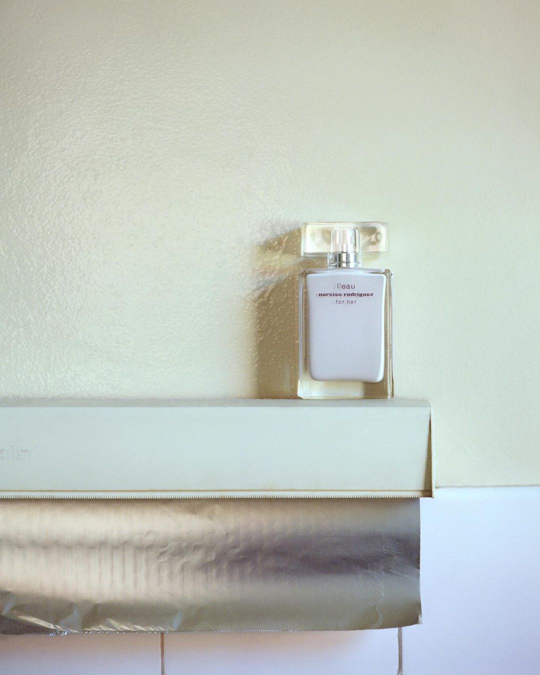 A bottle of white eau de parfum sits on a roll of foil in a brightly lit indoor setting, capturing a minimalist beauty scene.