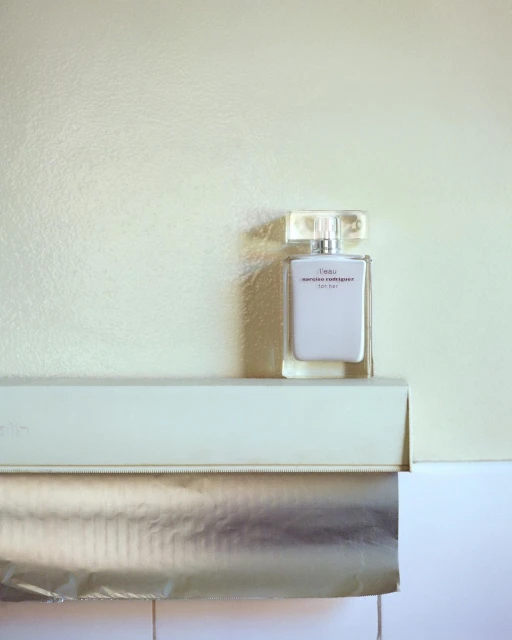 A bottle of white eau de parfum sits on a roll of foil in a brightly lit indoor setting, capturing a minimalist beauty scene.