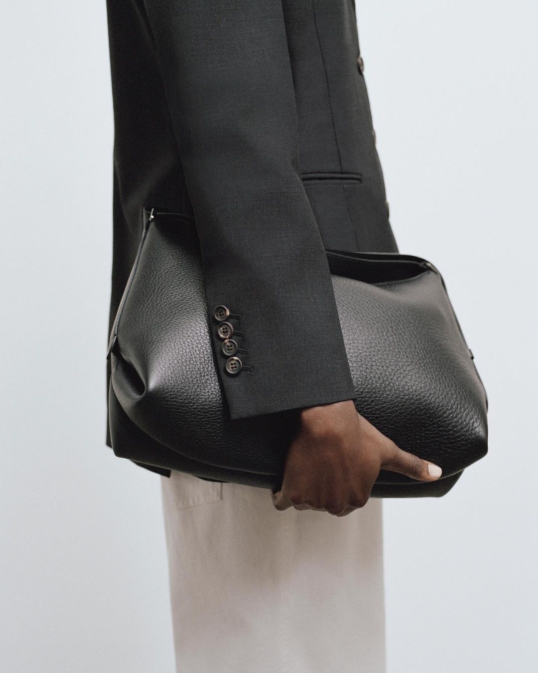 A person in a dark blazer holds a black leather shoulder bag in a studio setting, presenting a sophisticated fashion statement.