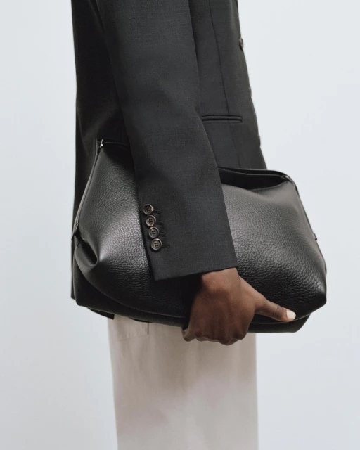 A person in a dark blazer holds a black leather shoulder bag in a studio setting, presenting a sophisticated fashion statement.