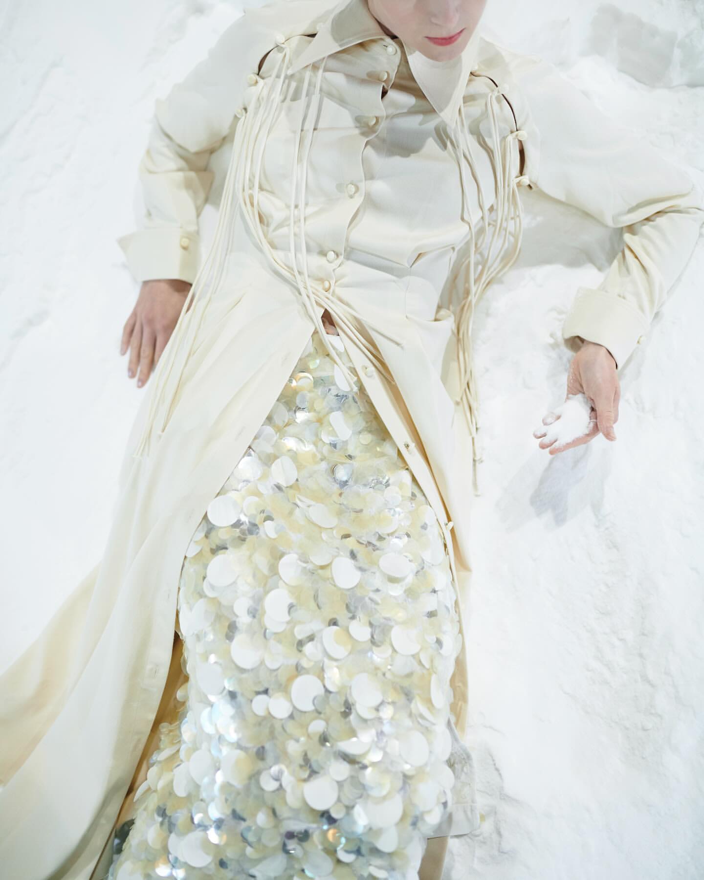 Adult female in cream-colored coat and sequin skirt lays on white surface. Soft lighting, shallow depth-of-field in studio setting.