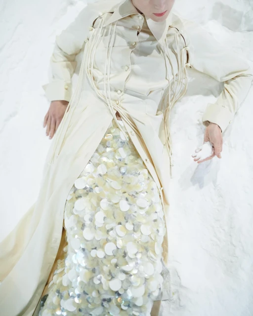 Adult female in cream-colored coat and sequin skirt lays on white surface. Soft lighting, shallow depth-of-field in studio setting.