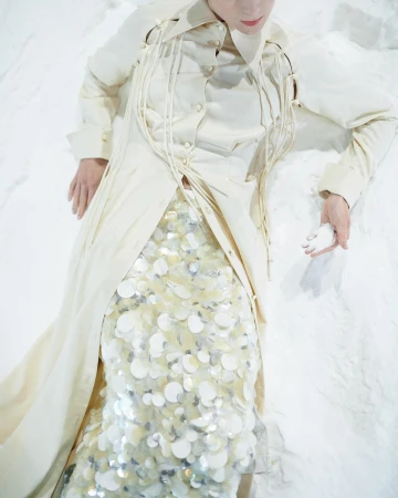 Adult female in cream-colored coat and sequin skirt lays on white surface. Soft lighting, shallow depth-of-field in studio setting.