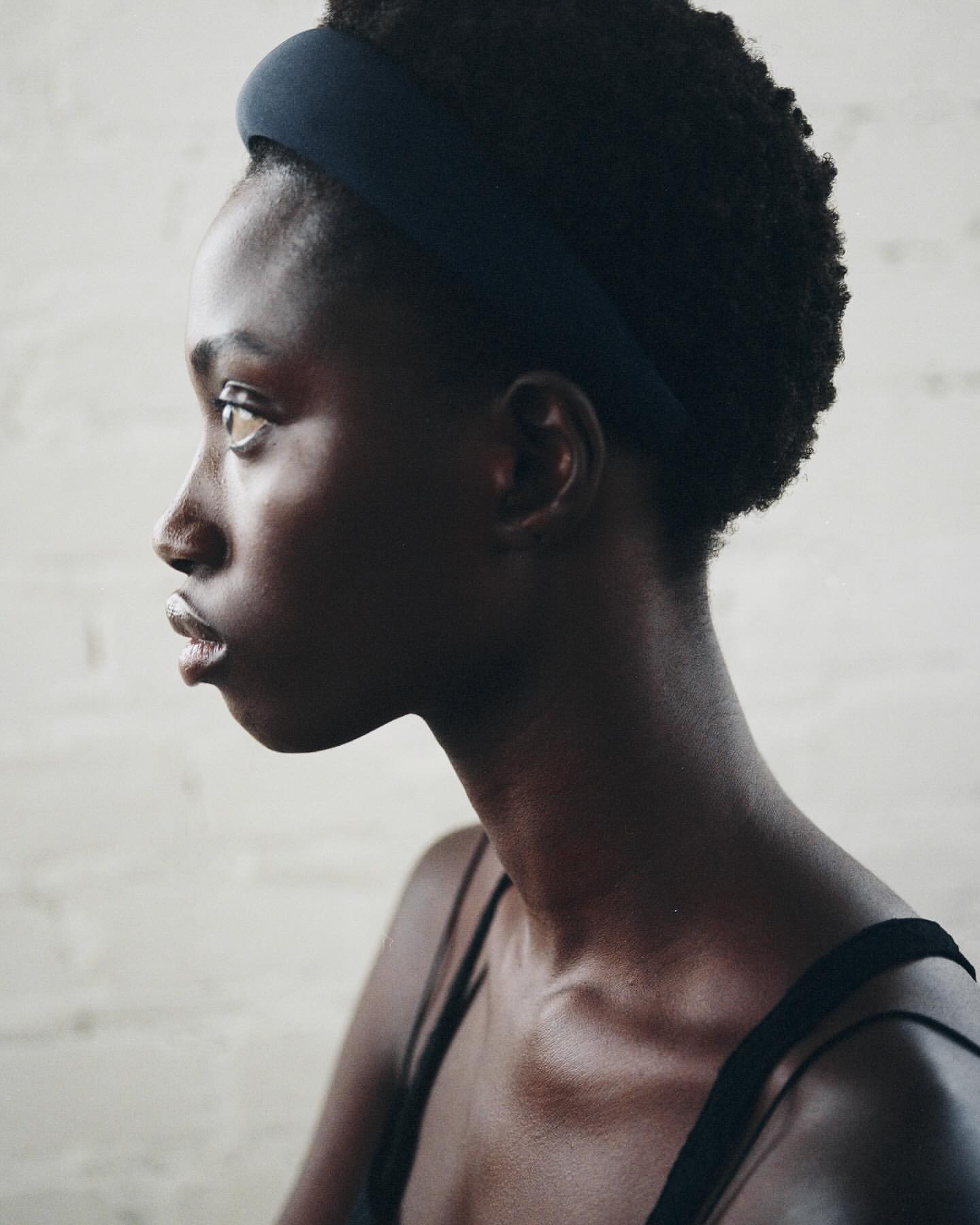 Profile shot of a Black woman with dark curly hair, wearing a headband and tank top against a textured neutral background.