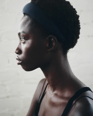 Profile shot of a Black woman with dark curly hair, wearing a headband and tank top against a textured neutral background.