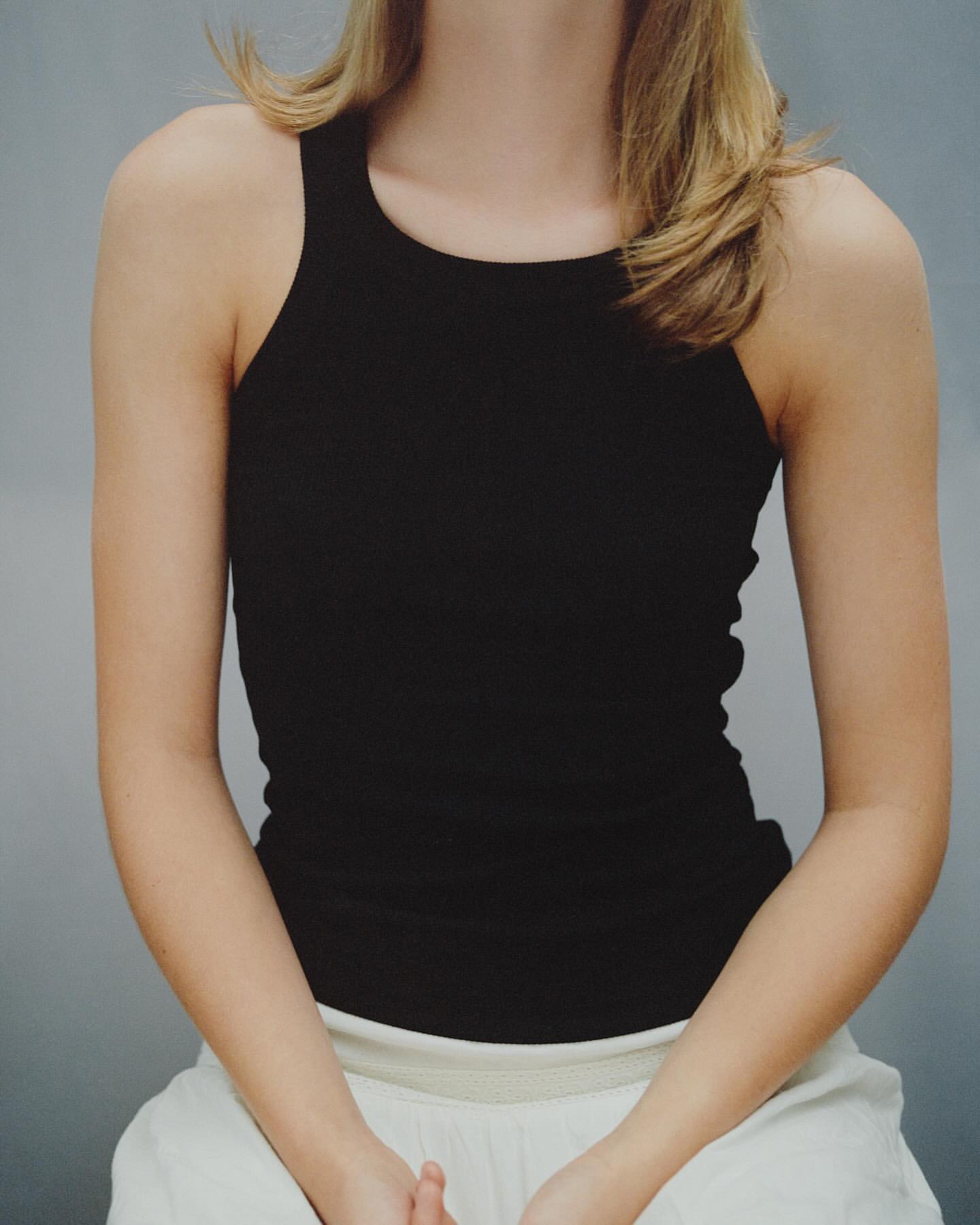 Close-up portrait of a woman wearing a black ribbed tank top against a plain grey seamless background.