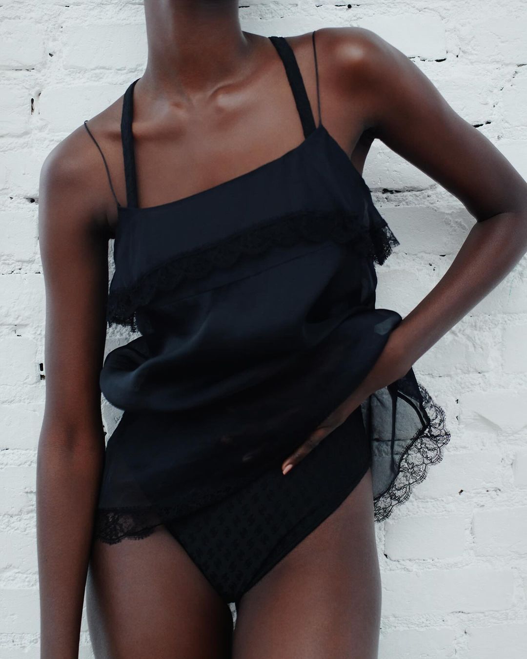A woman with dark skin poses in a black lace-trimmed camisole and underwear against a white brick wall in a studio setting.