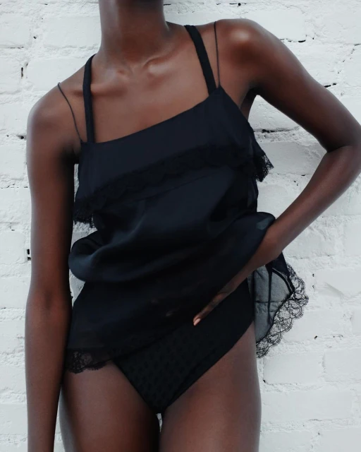 A woman with dark skin poses in a black lace-trimmed camisole and underwear against a white brick wall in a studio setting.