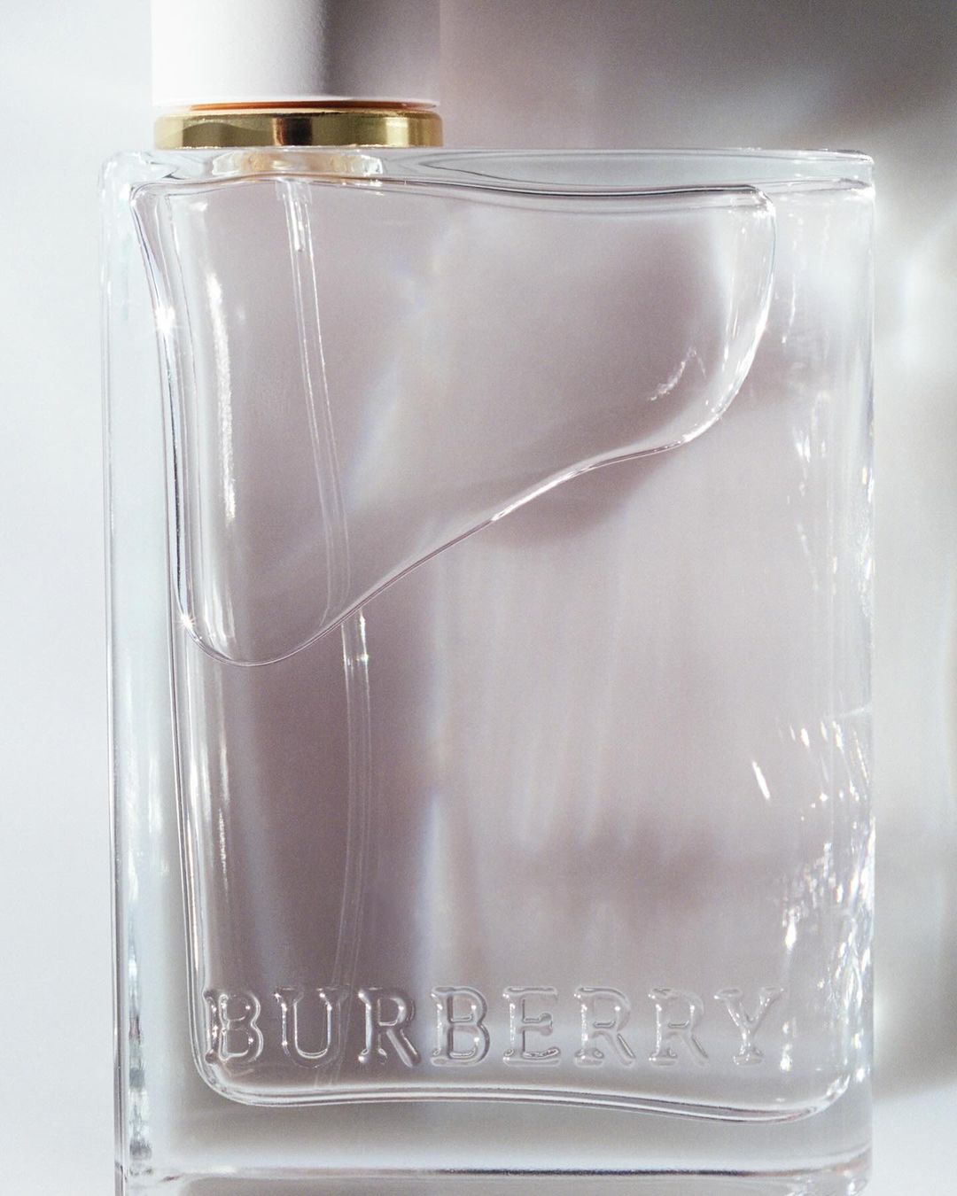 A clear glass Burberry fragrance bottle with a white cap and gold accent is set against a white background in a studio shot.