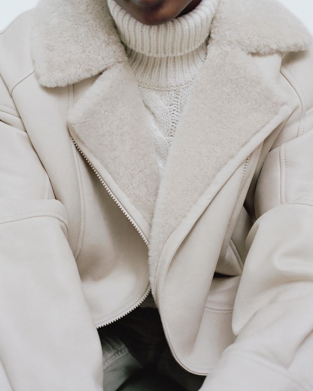 Close-up of a woman wearing an unzipped, ivory-colored, faux-shearling jacket over a high-neck knit sweater. Fashion shoot.