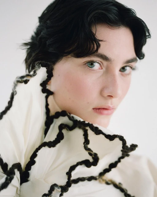 Close-up portrait of a dark-haired woman with pale skin wearing a cream-colored garment with black trim, against a white backdrop.