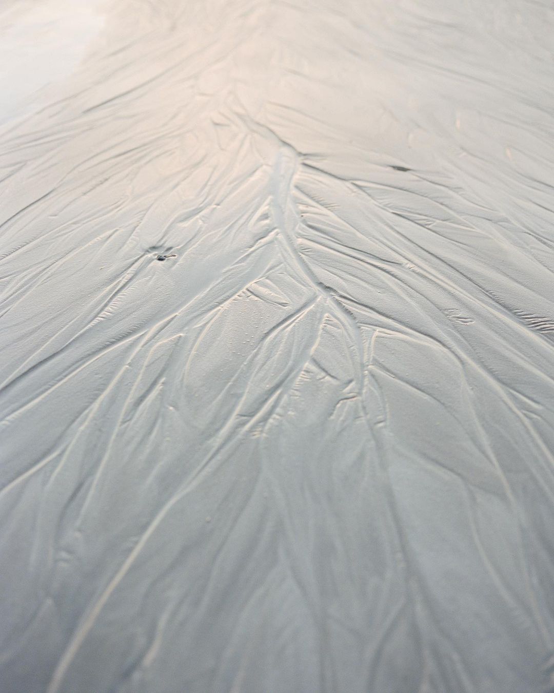 High-angle shot of white, sandy beach surface with winding water marks creating a textured, natural pattern that leads to the horizon.