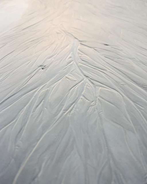 High-angle shot of white, sandy beach surface with winding water marks creating a textured, natural pattern that leads to the horizon.