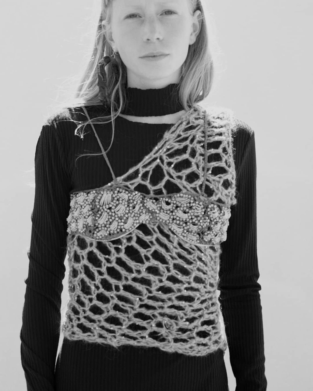 Monochrome portrait of a fair-skinned model with long blonde hair wearing an embellished knit top and black long-sleeve shirt against a white background.