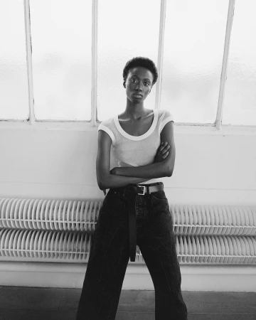 Black woman in a short-sleeve top and dark trousers posing in front of a window and radiator in Paris by Aude Le Barbey