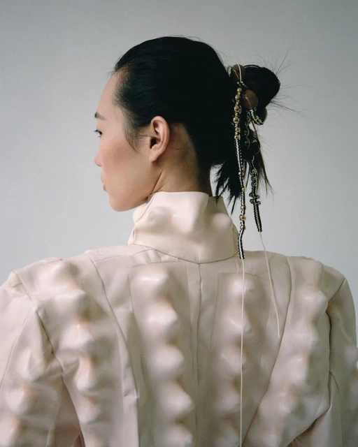 Back view of a fashionable adult woman with dark hair, wearing a light pink high-collar textured jacket in a minimalist studio.