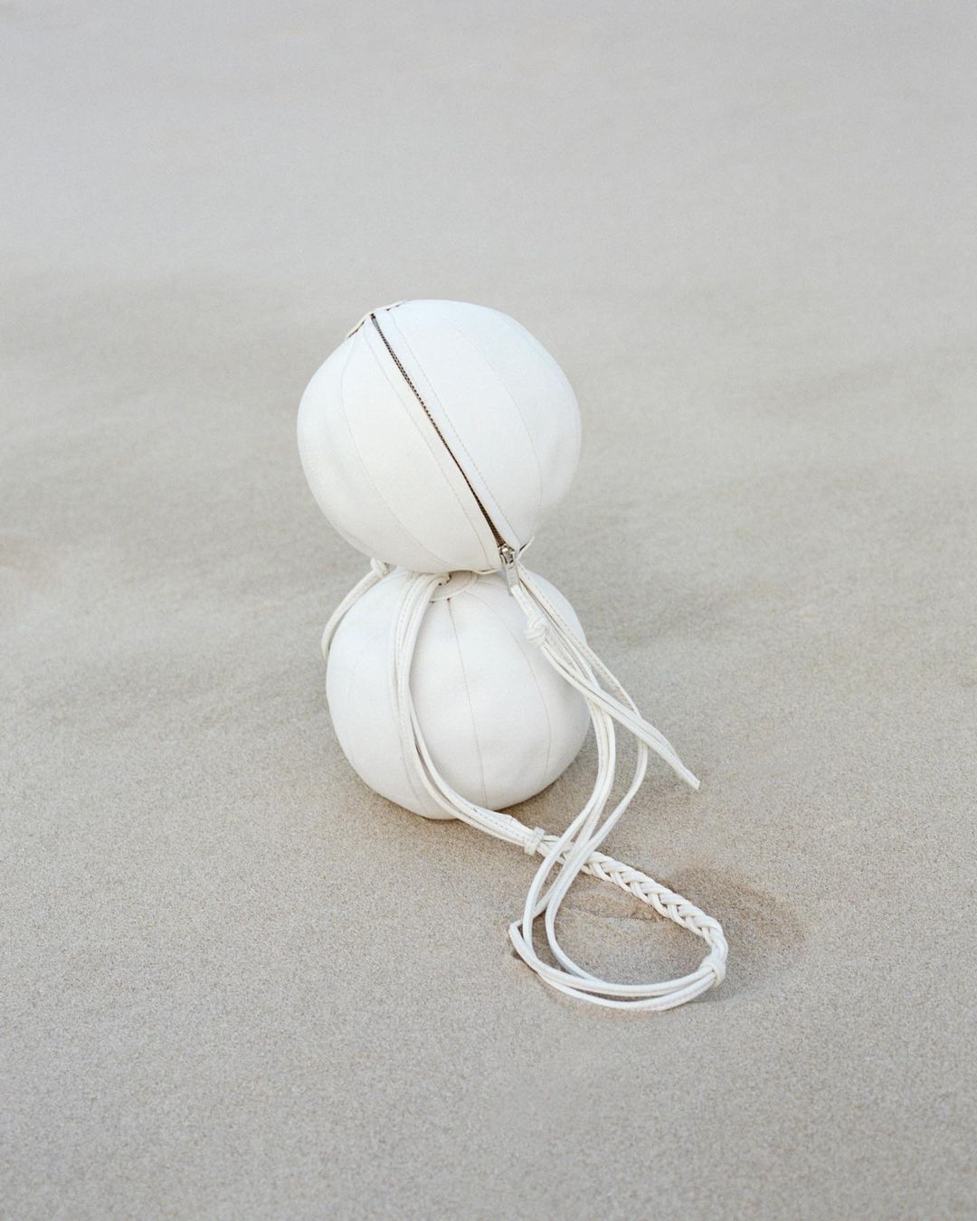 Two stacked white spherical bags with braided cords on a neutral surface. The minimalist image emphasizes texture and round shapes.