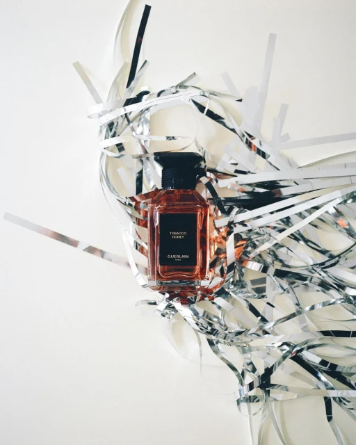 Perfume bottle of Tobacco Honey by Guerlain surrounded by shiny silver streamers on a white surface in a beauty shot.