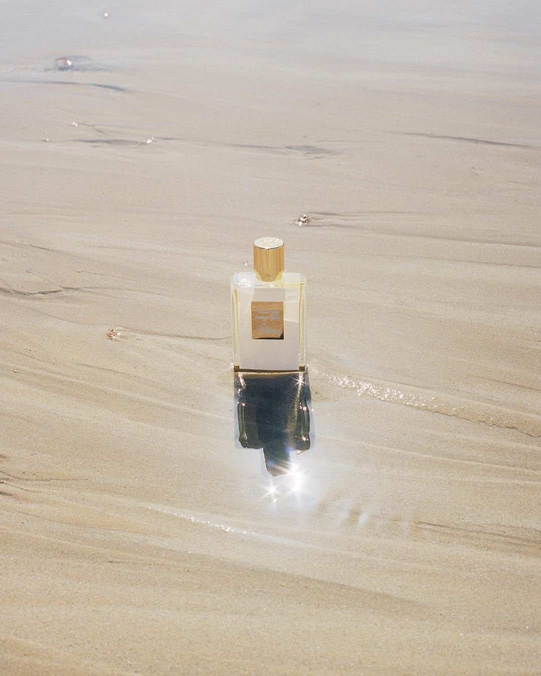 A bottle of clear glass perfume with a gold cap stands upright on a sandy beach reflecting in the shallow water.