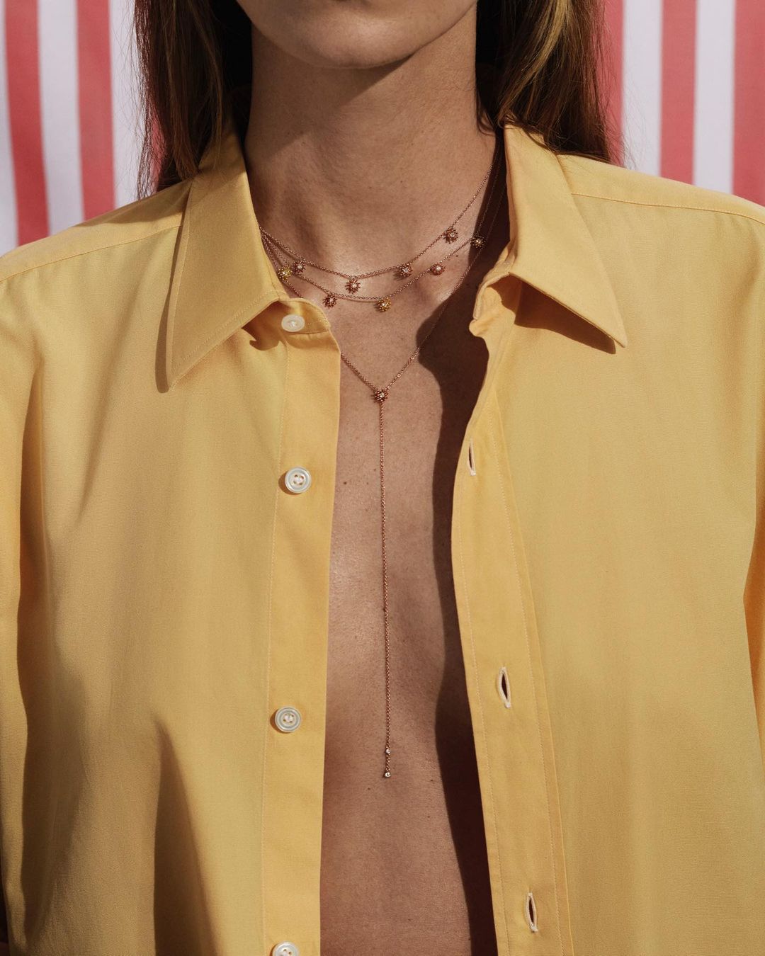 A woman in a bright yellow shirt poses with three layered necklaces in front of a red and white striped background, close-up shot.
